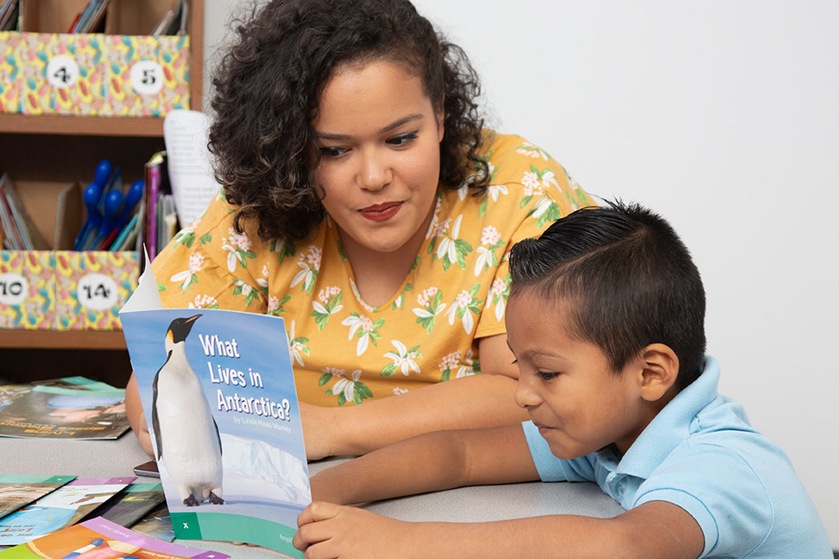 A teacher watching a little boy reading a book about penguins